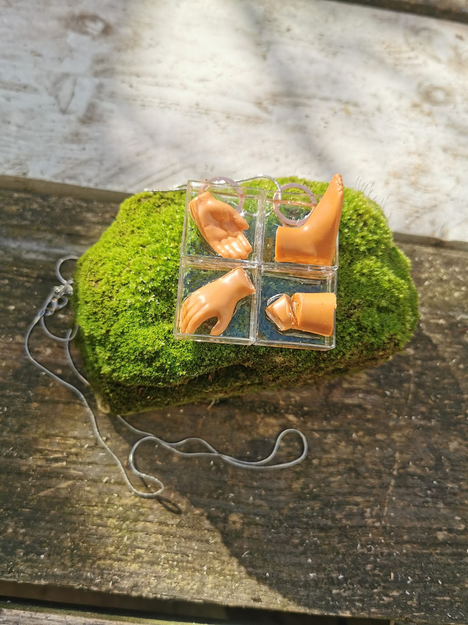 Hanger aan ketting: doorzichtig kubusje met poppenhandjes, gefotografeerd op mos
